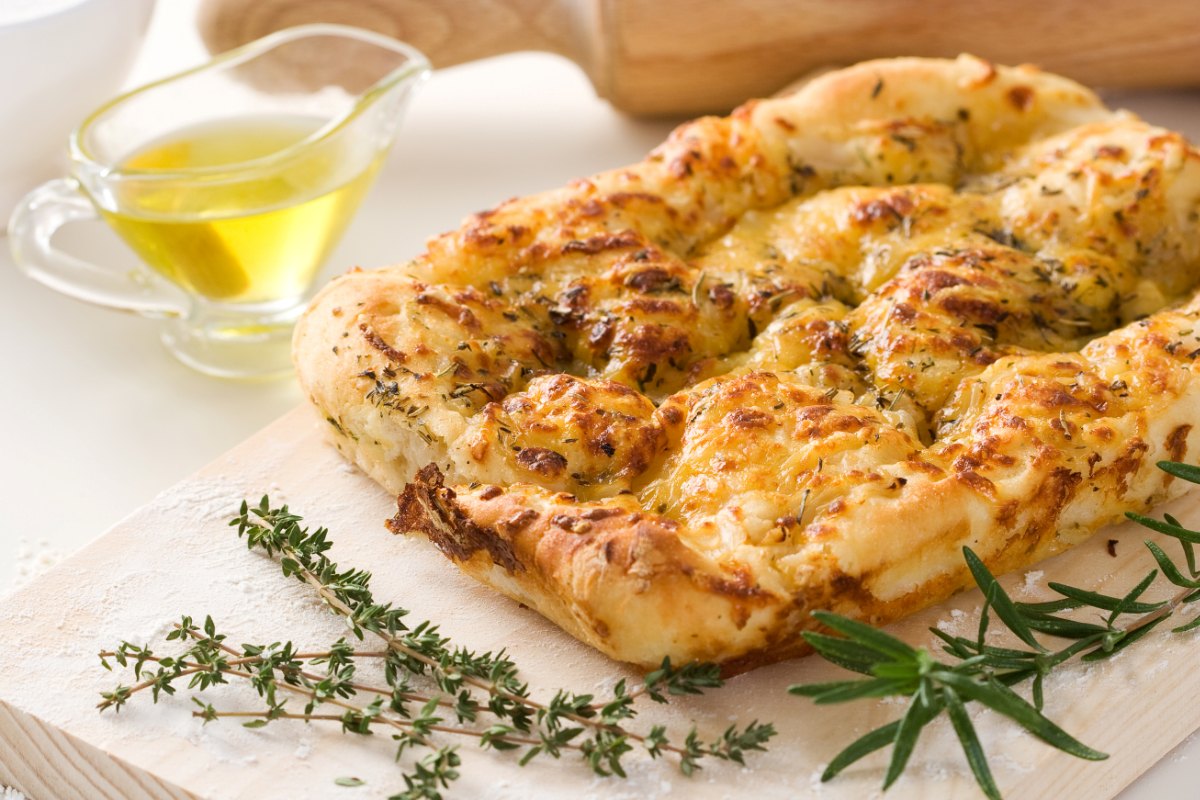 Focaccia z pieca na jasnej, drewnianej desce w towarzystwie świeżych ziół i oliwy