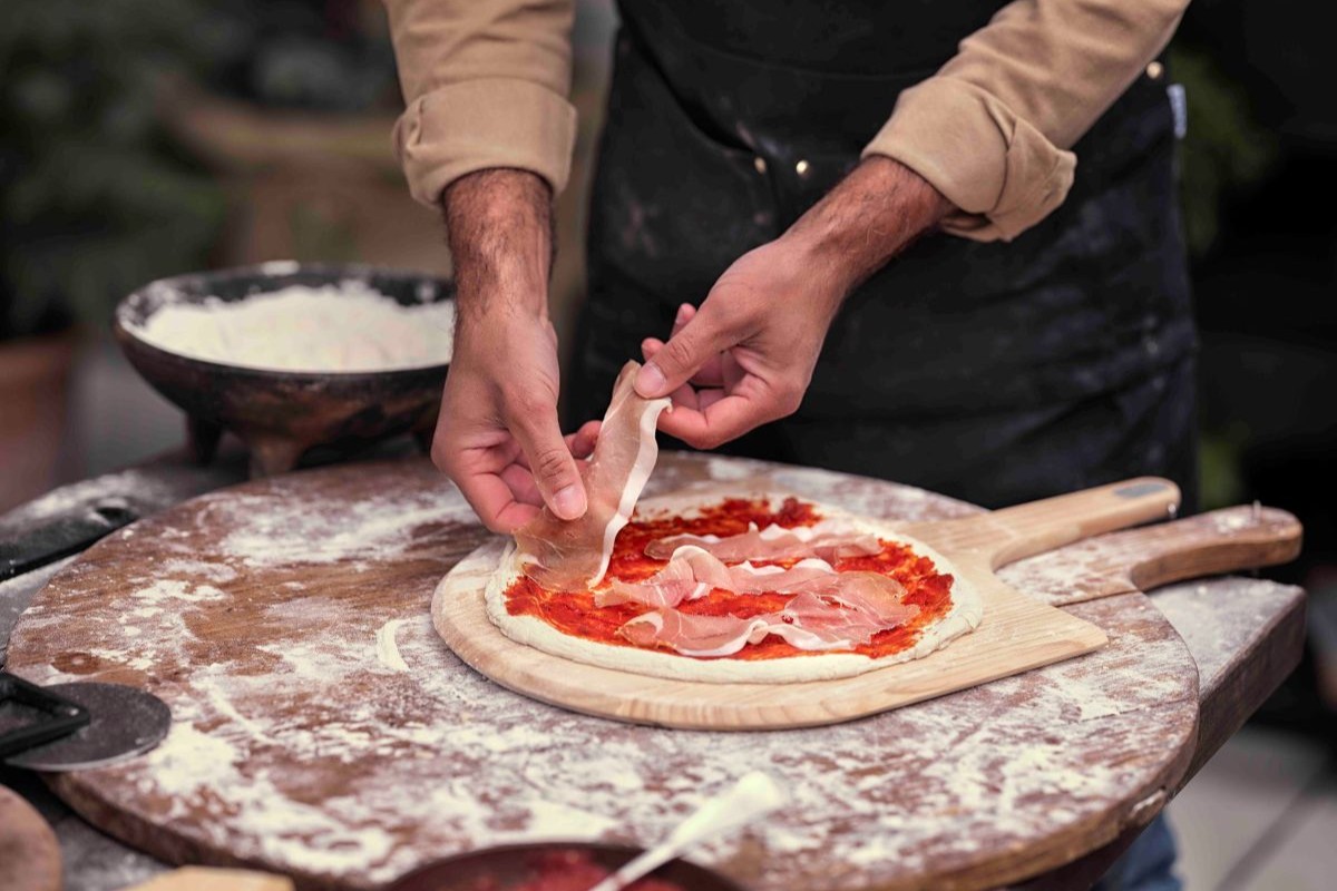 Męskie ręce kładą szynkę na pizzy gotowej do pieczenia, która leży na drewnianej łopacie
