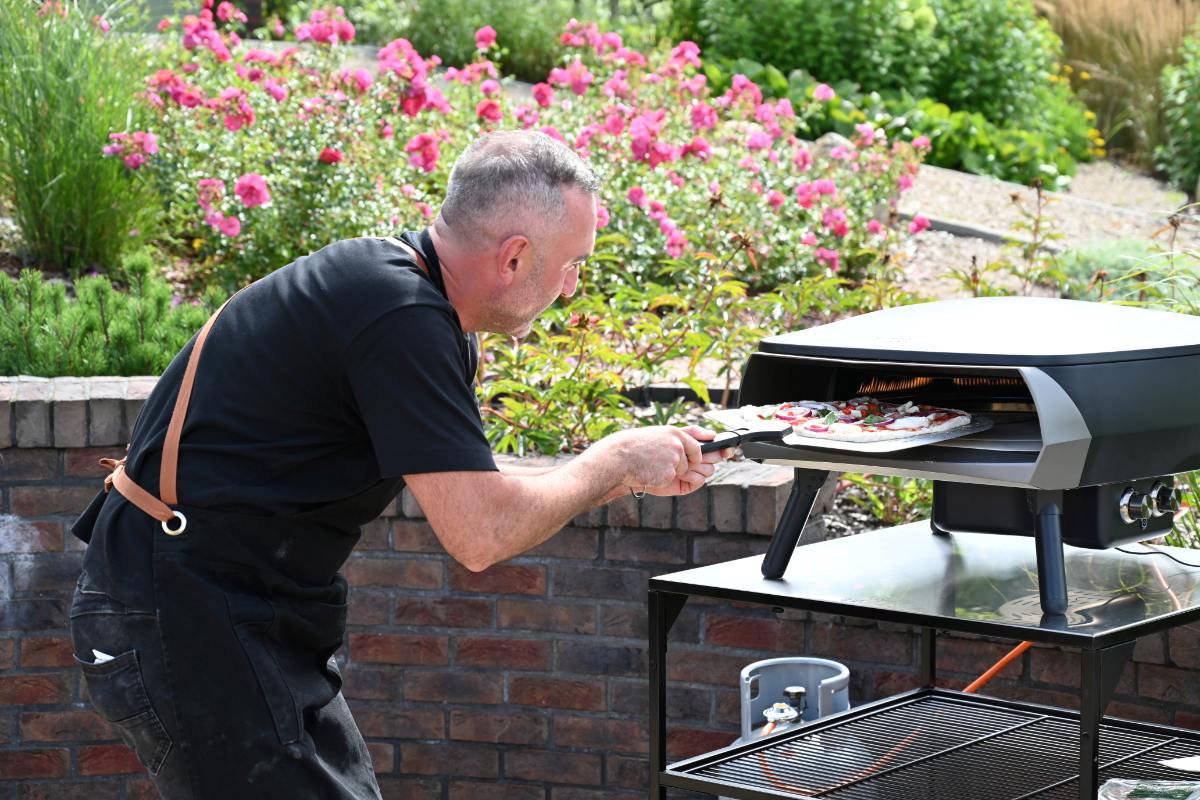 Scena w kwiecistym ogrodzie. Pizzaiolo zagląda do pieca Witt sprawdzając wypieczenie pizzy