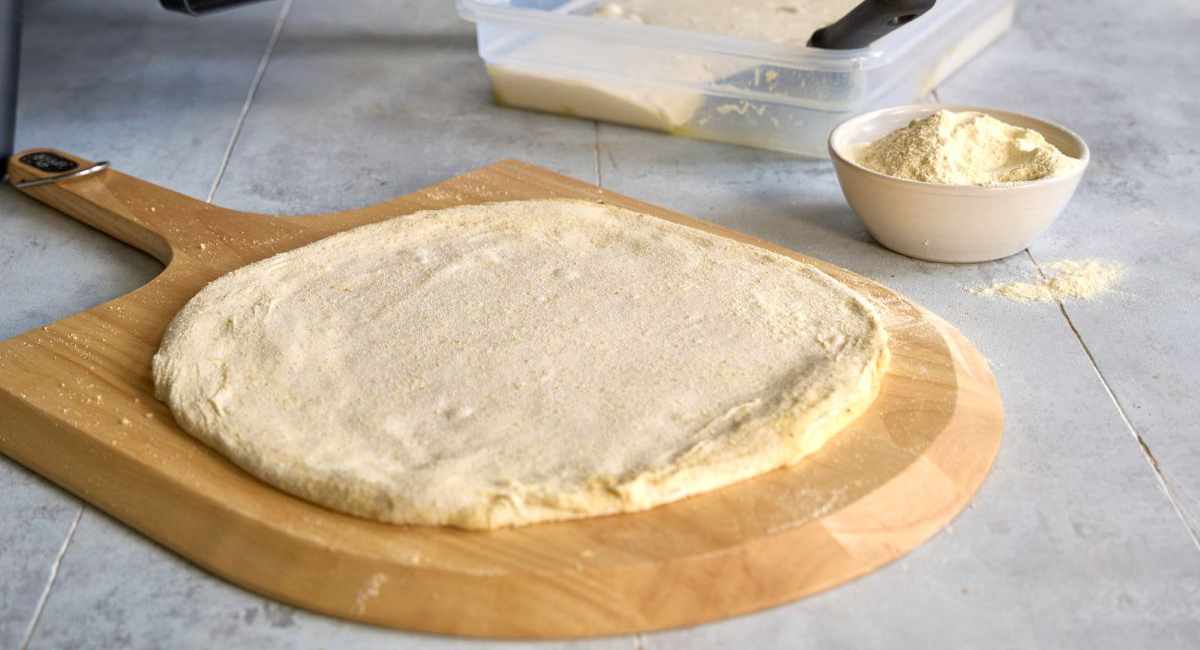Rozciągnięte ciasto na pizzę leży na drewnianej desce. W tle pojemnik z kulkami na pizzę