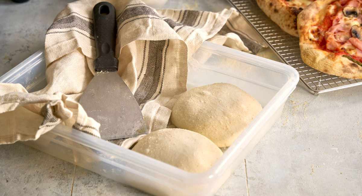 Kulki ciasta na pizzę w pojemniku pod ściereczką i szpachelka do pizzy. W tle na kratce studzi się pizza