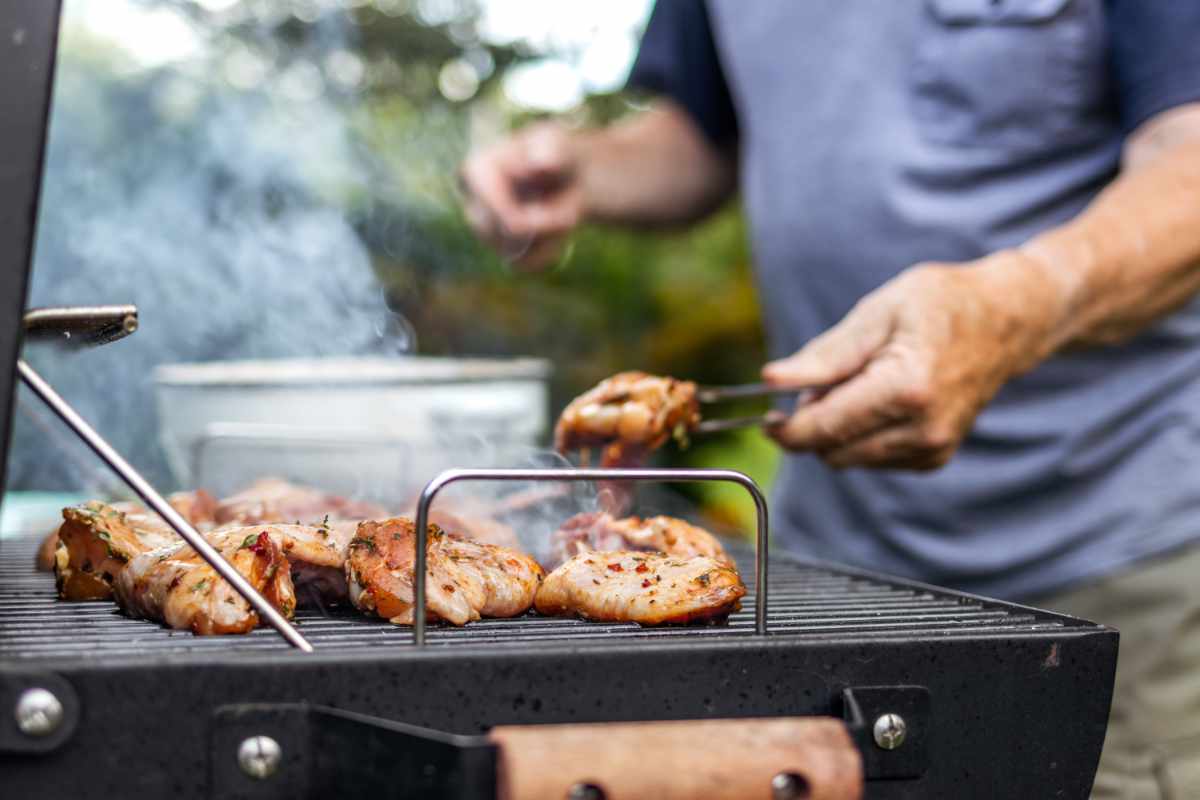 Mężczyzna w niebieskiej koszulce obraca kawałki mięsa na ogrodowym grillu.