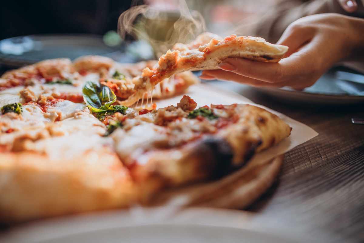 Świeżo wypieczona pizza i dłoń sięgająca po kawałek z ciągnącym się serem