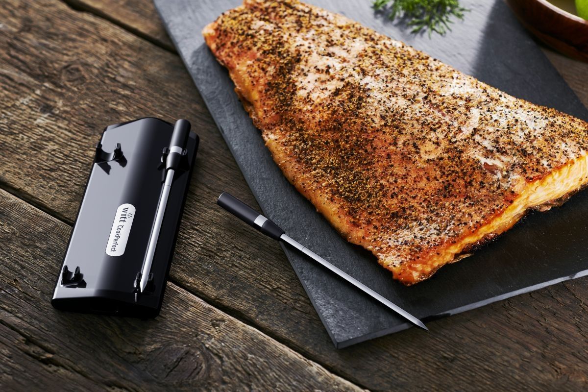 Pieczony łosoś na kamiennej podstawce z wbitym termometrem CookPerfect. Obok leży baza CookPerfect.